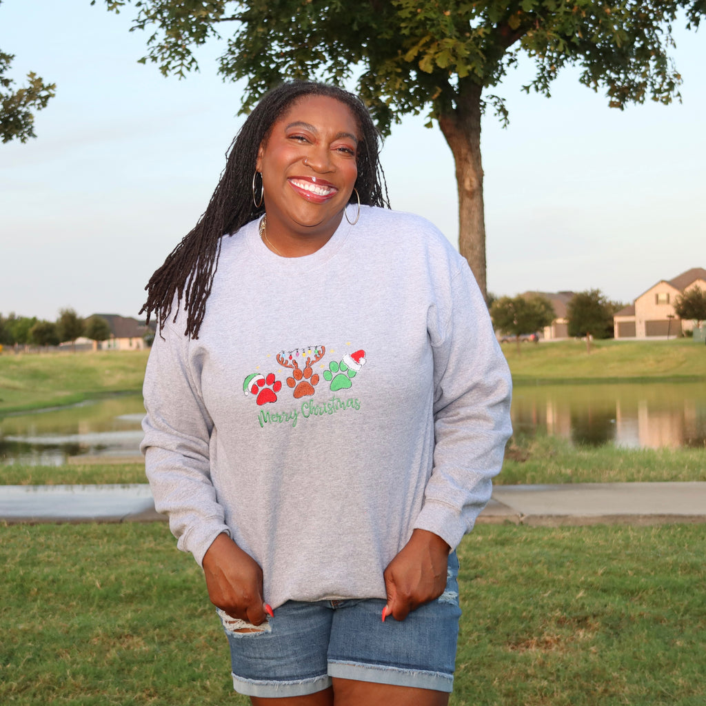 A smiling woman stands outdoors in a grassy park area wearing a light gray crewneck sweatshirt with a Christmas-themed graphic and 'Merry Christmas' text, paired with denim shorts.